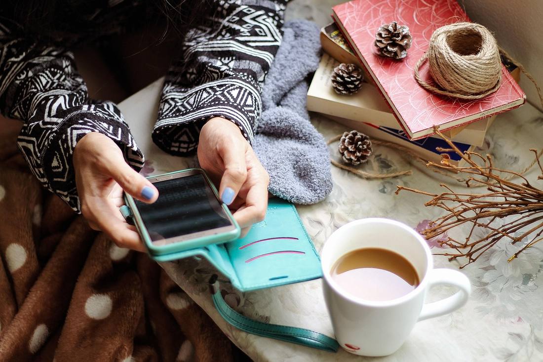 A person holding a smartphone in a teal case over a table with a cup of coffee, books and pine cones.