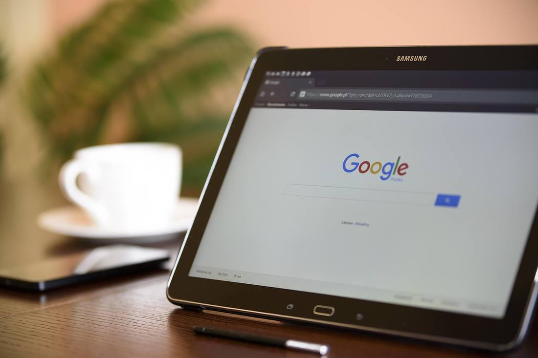 A Samsung tablet on a wooden desk showing the Google homepage in a web browser, next to a cup of coffee and a stylus.