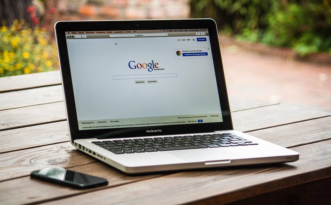 A MacBook Pro on a wooden table outdoors showing the Google homepage in a web browser, with a smartphone beside it