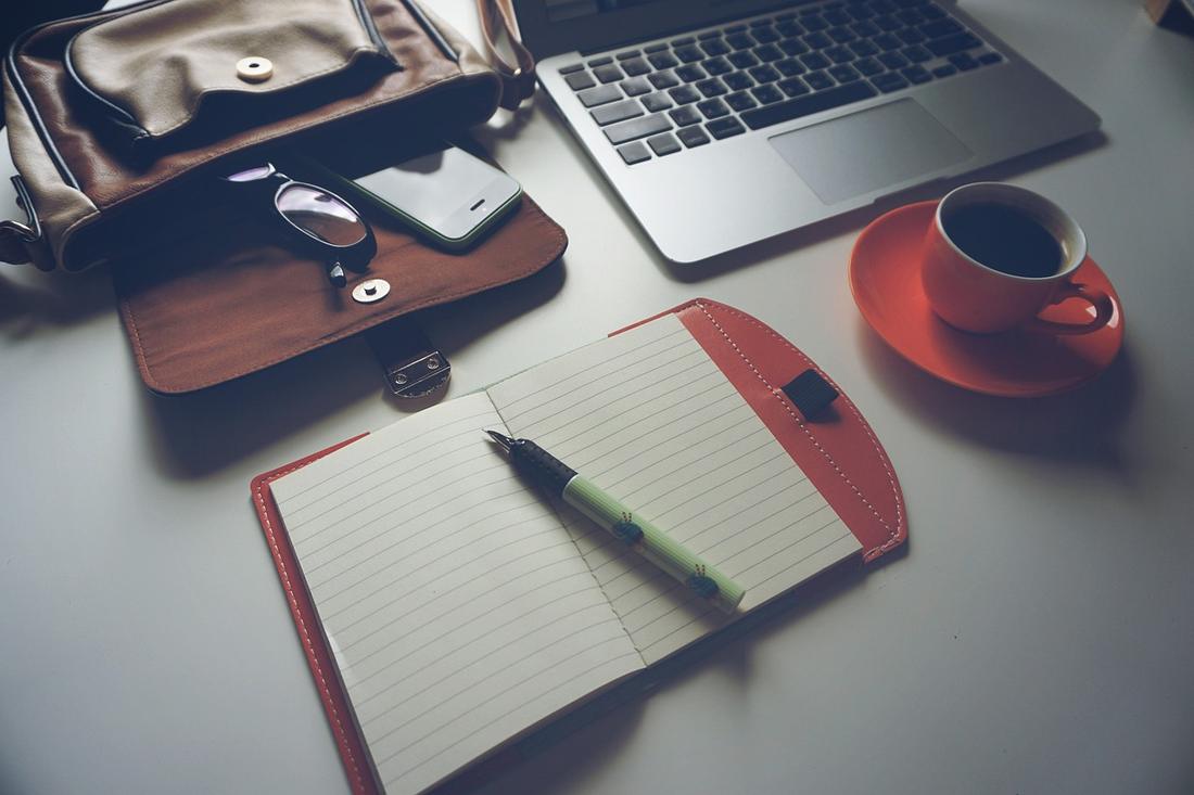 An open blank notebook with a pen on a desk, beside a laptop, a red cup of coffee, and a bag holding glasses and a phone.