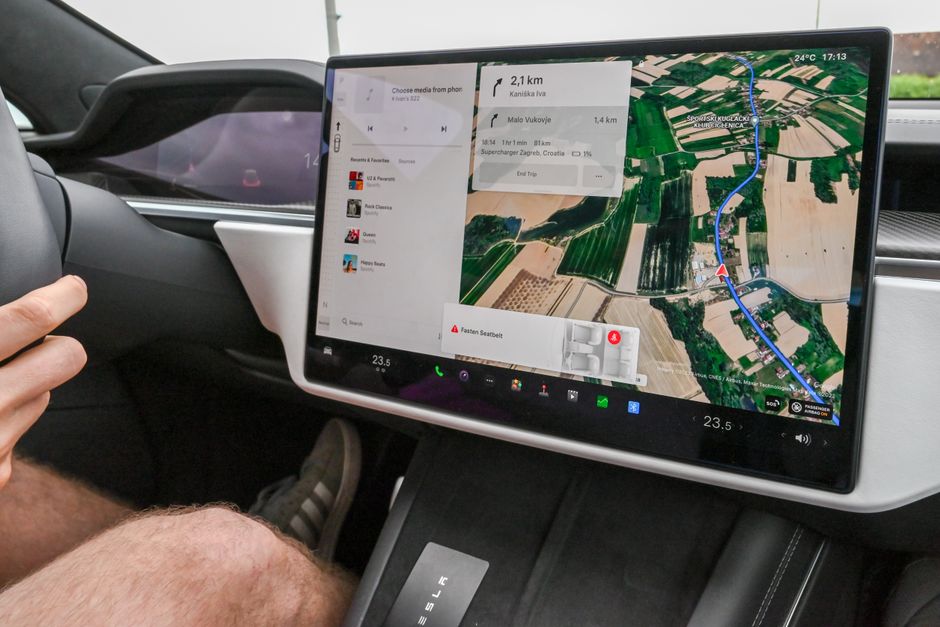 A large landscape touchscreen in a Tesla showing satellite navigation over farmland in Croatia, a media panel listing Spotify playlists, a Fasten Seatbelt warning and a cabin temperature of 23.5