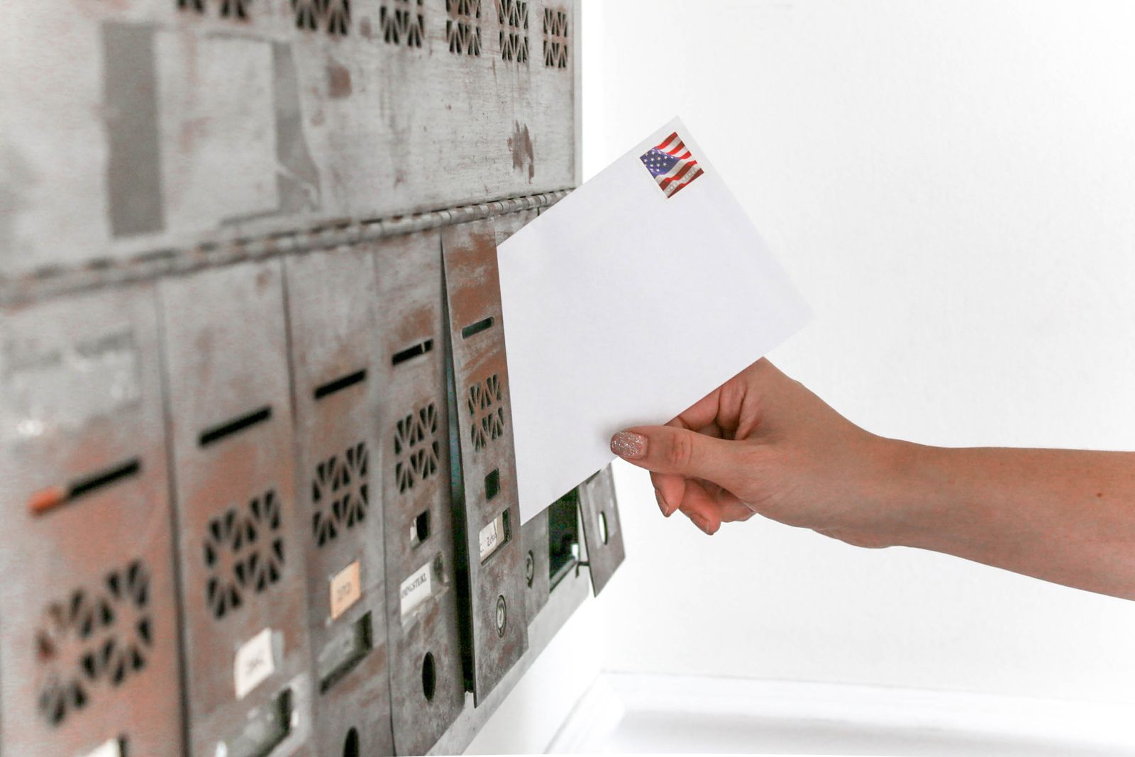 A hand posting a plain white envelope with a stamp into one of a row of grey metal apartment letterboxes, several of the slots below it still holding mail.