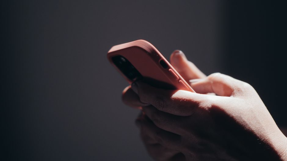 A hand holding a phone in a salmon-pink case, seen from behind against a dark background with warm light along its edge