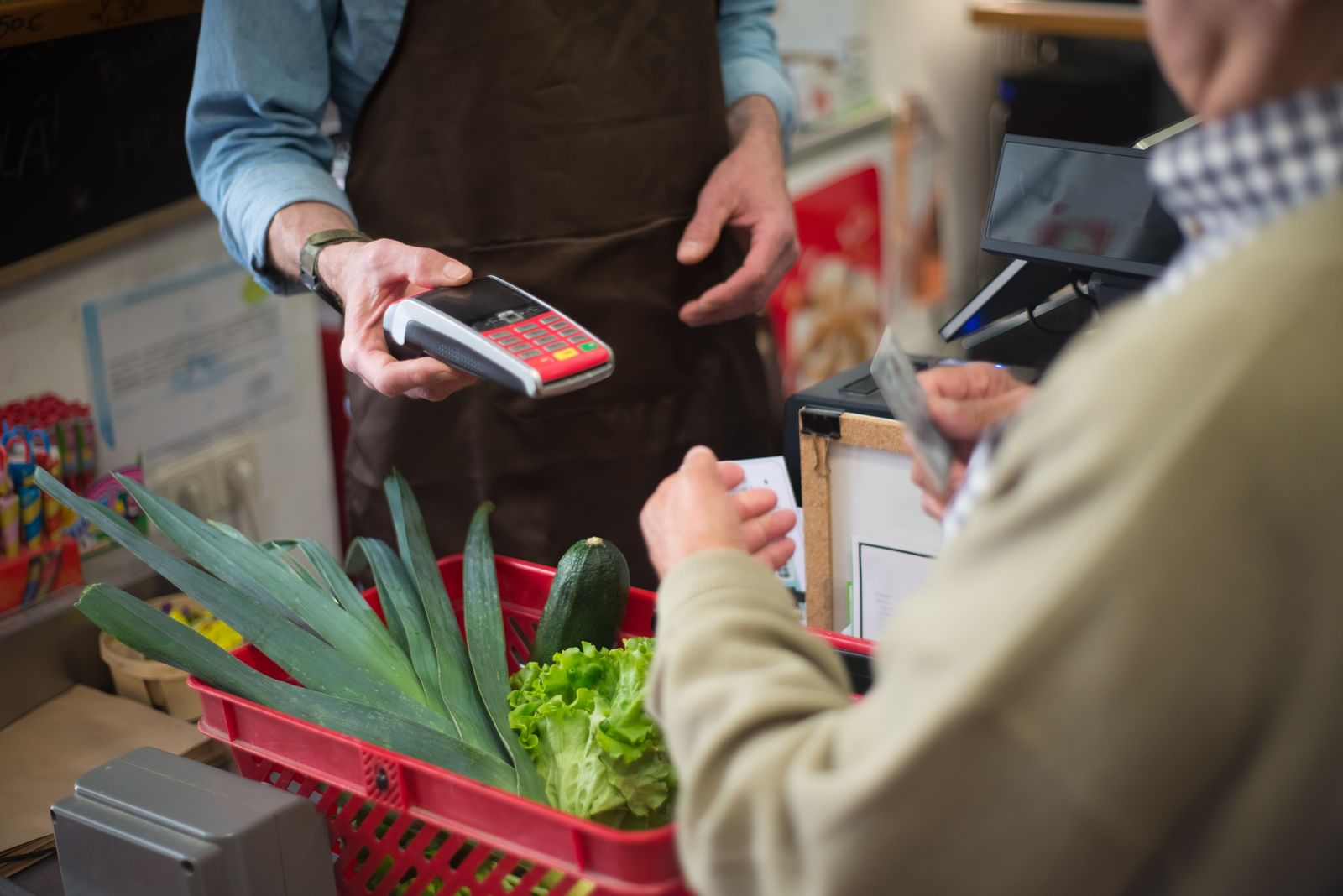 En la caja, un comerciante acerca un terminal de pago con tarjeta a un cliente que saca su tarjeta, con una cesta de verduras delante — la forma en que pagas es una de las señales más fáciles de vincular con tus hábitos de compra.