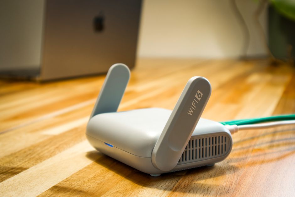 A small white WiFi 6 router with two raised antennas on a wooden floor, a green cable plugged into its side and a laptop out of focus behind it