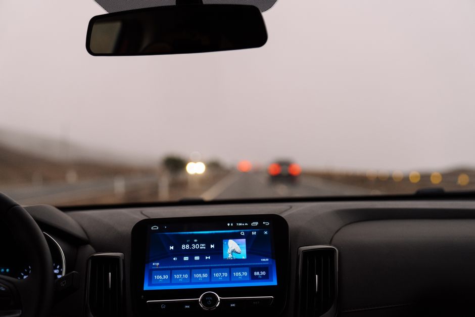 View through a car windscreen at dusk from the driver position: rear-view mirror above, a two-lane road in haze ahead with red tail lights, and a blue touchscreen showing FM radio on the dark dashboard