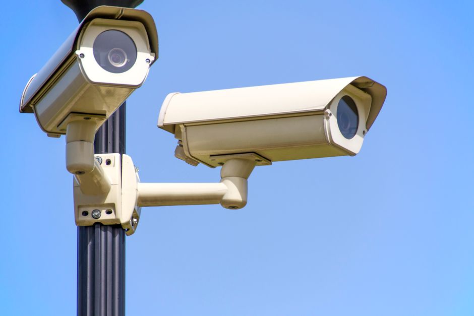 Two cream-coloured box cameras mounted on a black pole, photographed from below against a cloudless blue sky, one pointing left and one pointing right