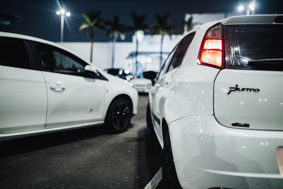 Two white cars parked side by side at night, shot low and close; the rear of the right-hand car shows a Punto badge and a lit red tail light, with palm trees and street lamps blurred behind.