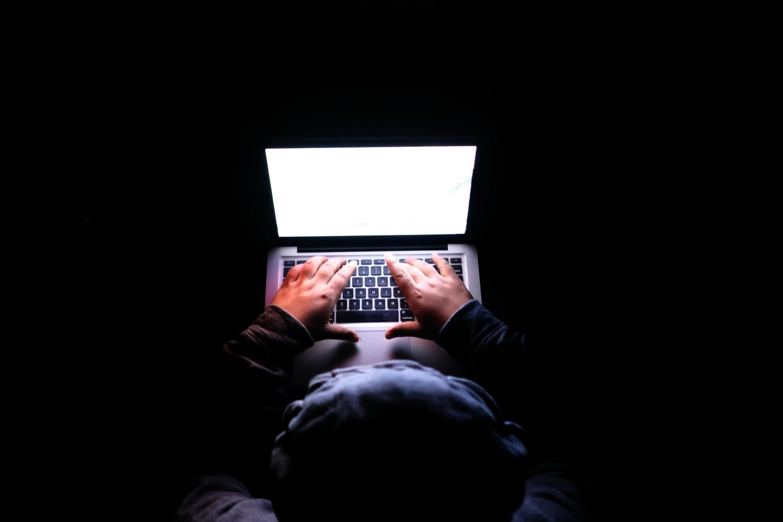 Une personne assise dans une pièce sombre, les mains sur le clavier d'un ordinateur portable vivement éclairé — la seule lumière visible venant de l'écran.