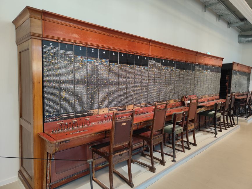 A manual telephone exchange preserved in a museum: a long wooden panel with rows of jacks numbered 0 to 9 and beyond, a line of operators' chairs still pushed up to it. Before any of this was digital, connecting two people already meant recording who called whom, and when.