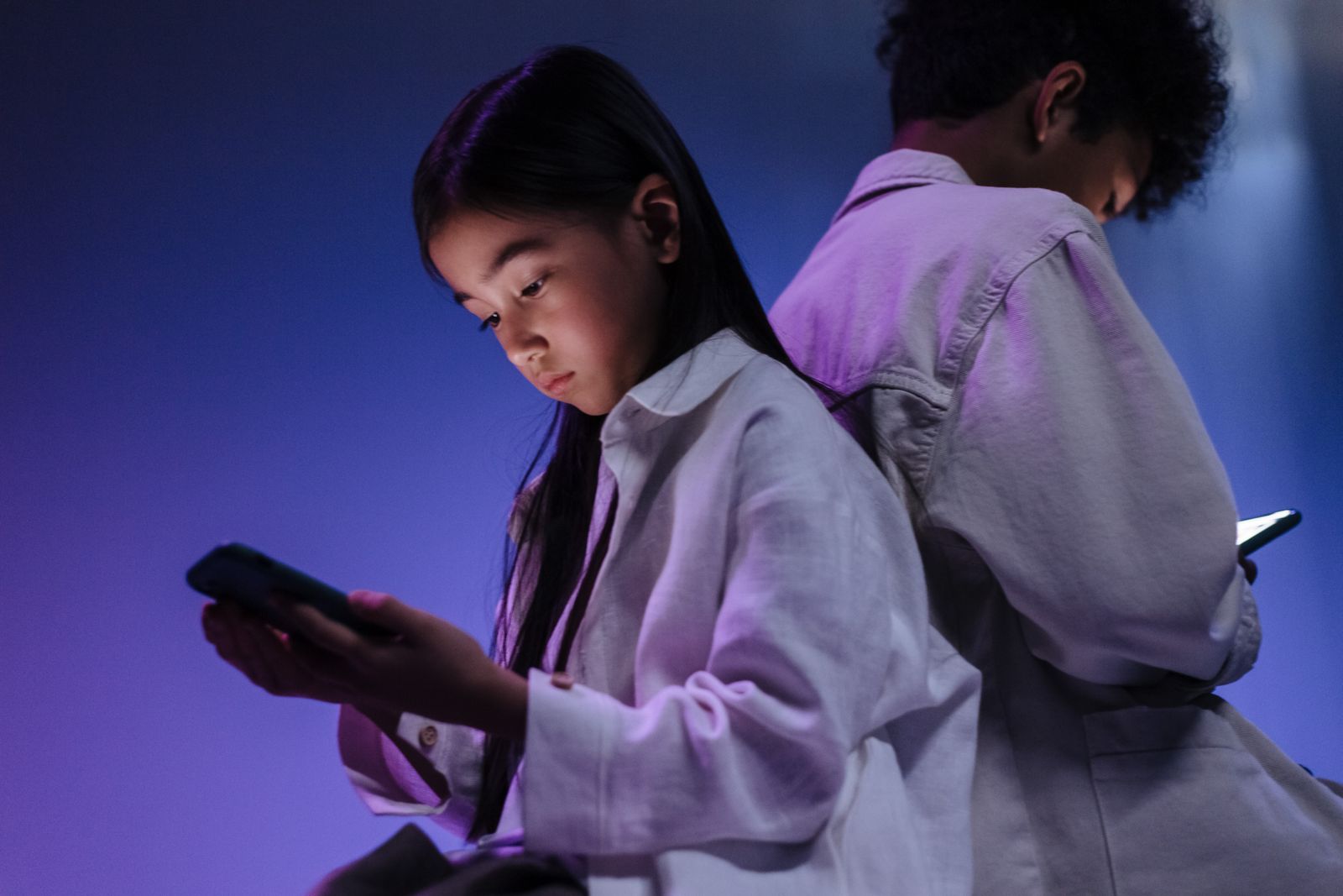 Two children sitting side by side, each absorbed in their own smartphone in a dimly lit room