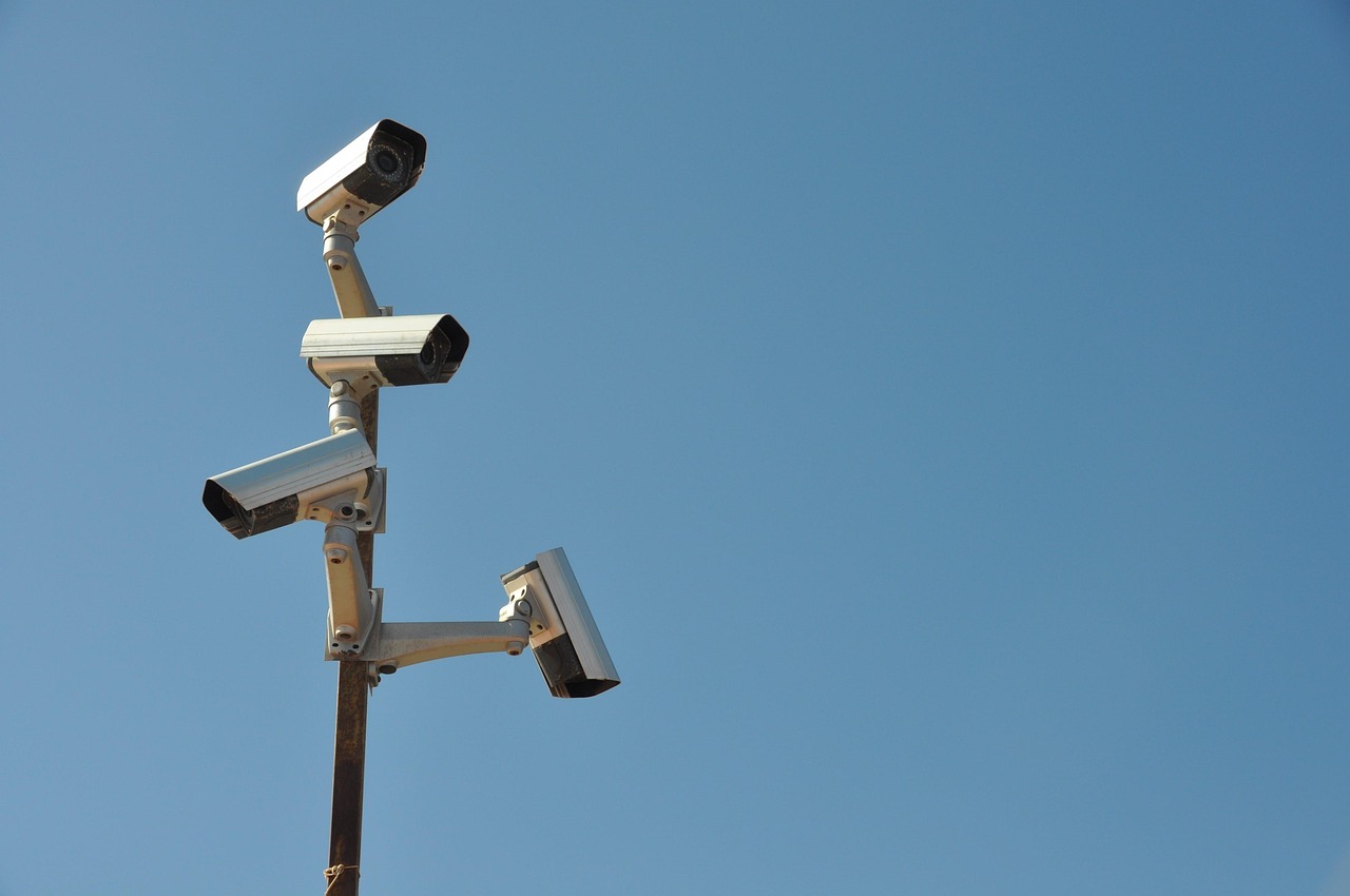 Four white surveillance cameras mounted at different heights on a single metal pole against a clear blue sky