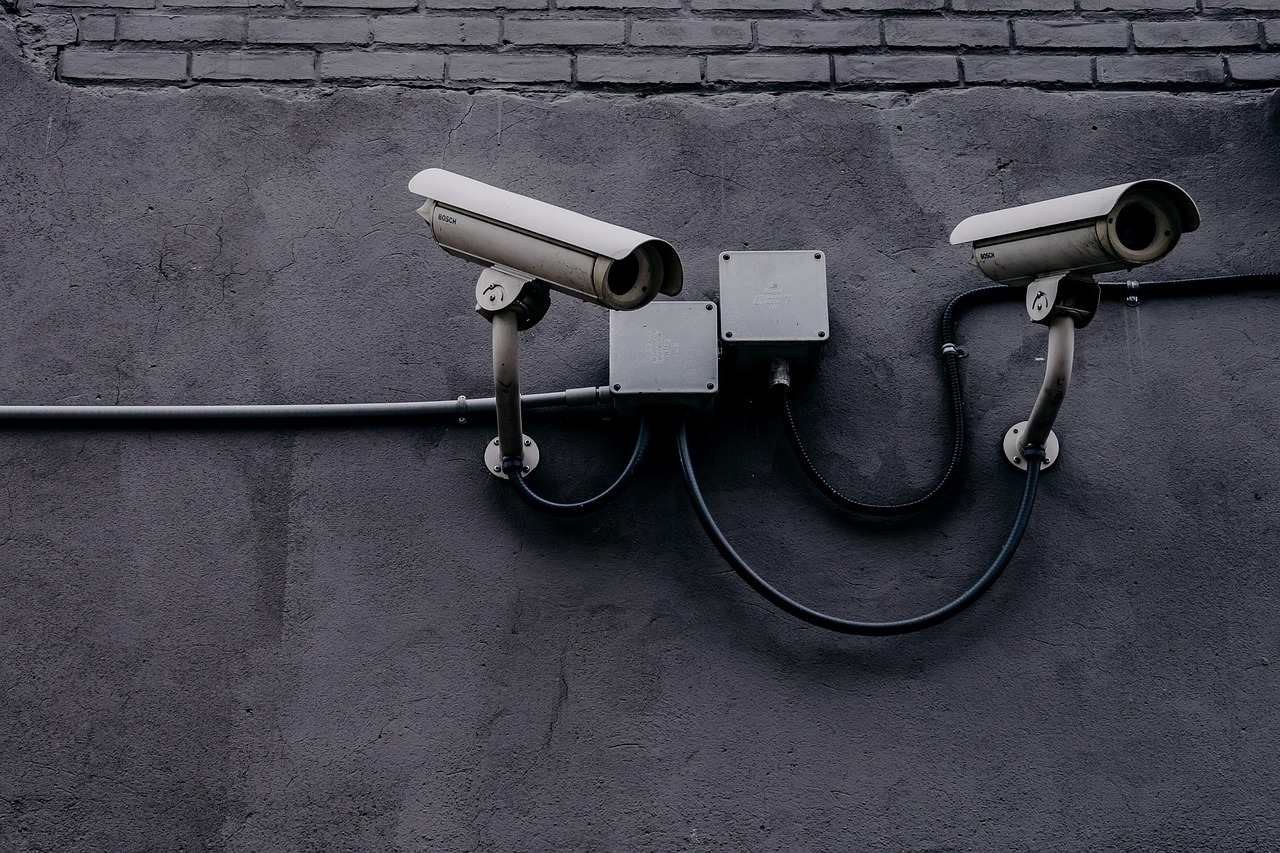 Two wired bullet CCTV cameras mounted on a grey painted brick wall, facing opposite directions and linked by conduit and junction boxes