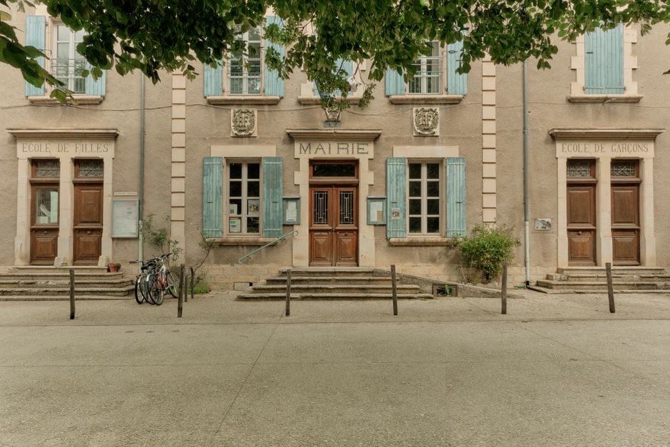 The facade of a small French town hall in beige stone, its shutters painted pale blue-green, MAIRIE carved above the central door and ÉCOLE DE FILLES and ÉCOLE DE GARÇONS above the side doors, with bicycles parked at the steps